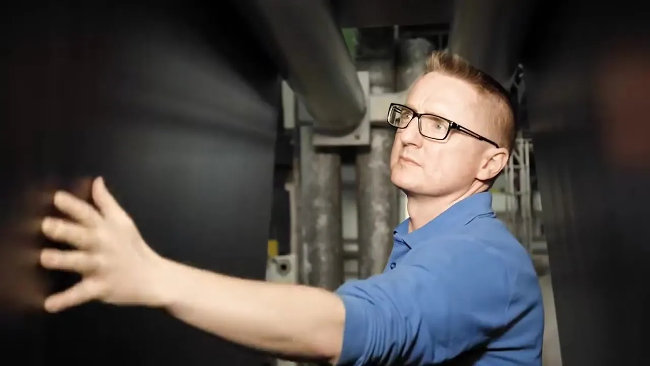 Filmstill: Mann mit Brille legt die Hand an eine Industrieanlage