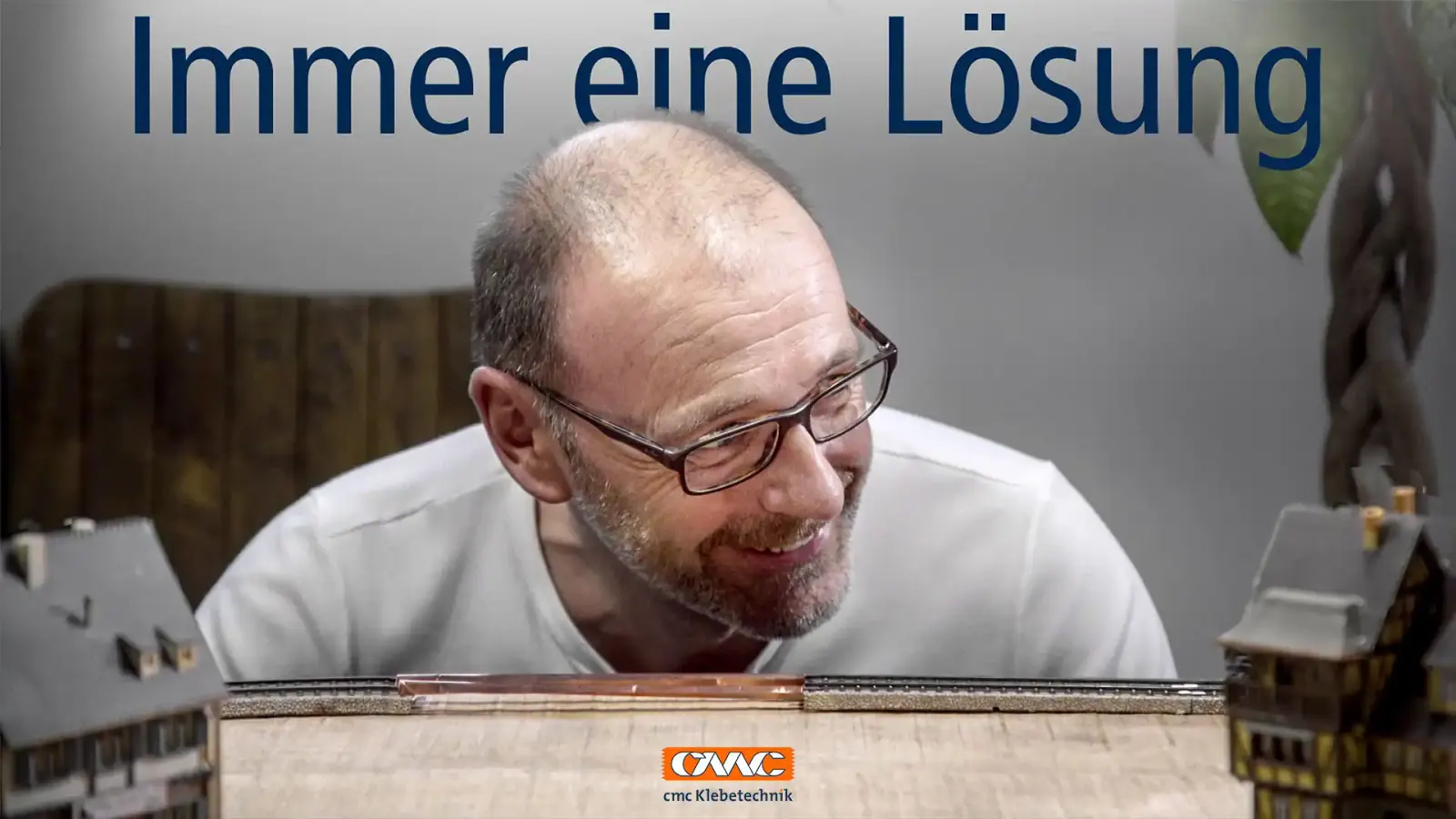 Mann mit Brille lächelt hinter einer Modelleisenbahn-Landschaft, darüber der Schriftzug „Immer eine Lösung“, Logo von CMC Klebetechnik