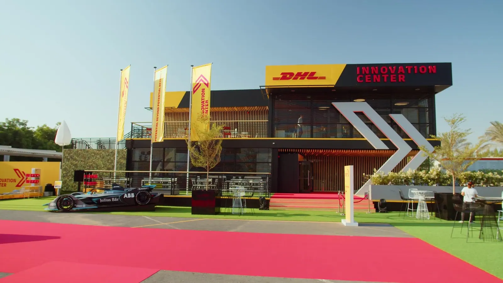 Filmbild aus „Innovation That Moves“: Gebäude des DHL Innovation Center mit gelben Fahnen, rotem Teppich und einem Rennwagen vor dem Eingang