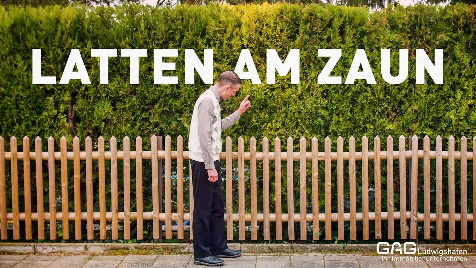 Mann zählt die Latten eines Holzzauns vor einer Hecke, Titel „Latten am Zaun“