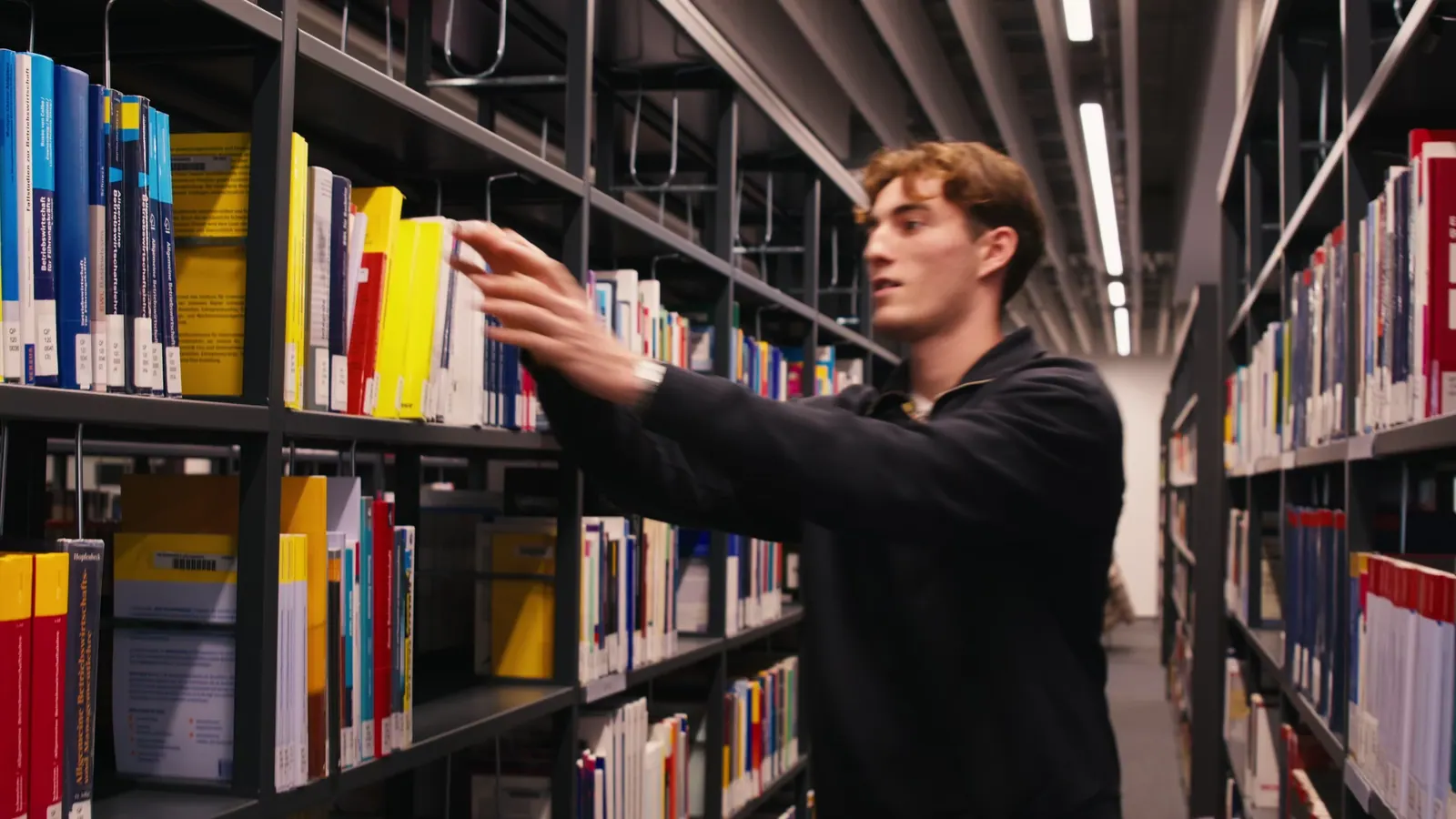 Student greift in einer Bibliothek nach einem Buch im Regal