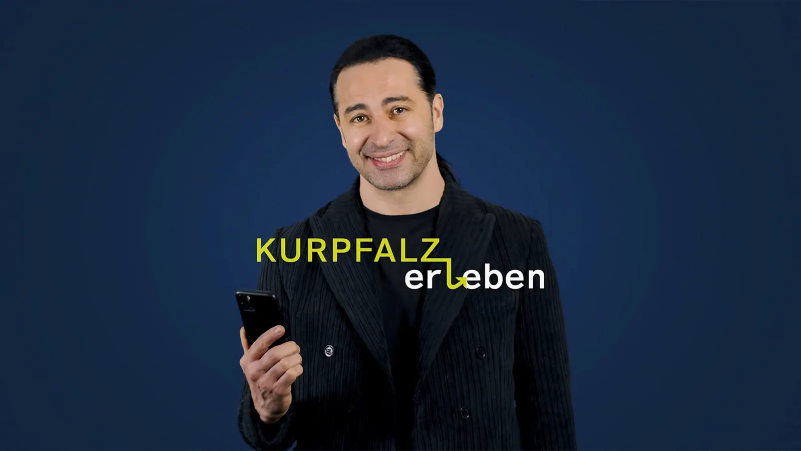 Mann im schwarzen Anzug hält ein Smartphone vor dunkelblauem Hintergrund, Schriftzug „KURPFALZ erleben“