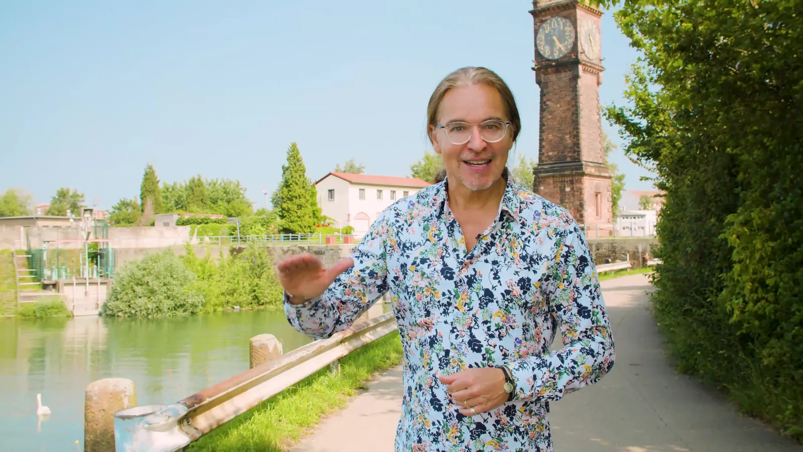 Mann im geblümten Hemd spricht am Kanalufer, dahinter ein Uhrturm