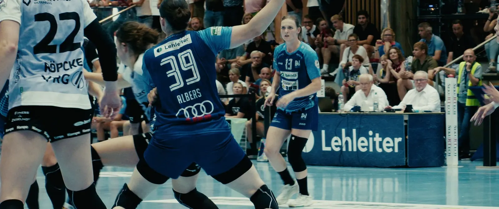 Handballszene in voller Halle: Spielerin mit der Nummer 39 im blauen Trikot zwischen Abwehrspielerinnen in Weiß