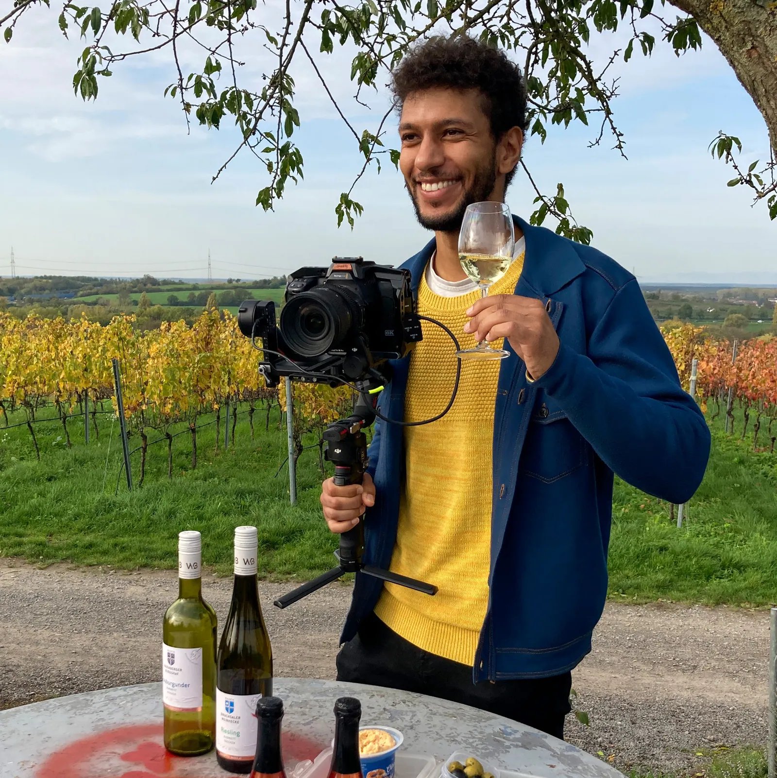 Hipolito Macamo mit Gimbal-Kamera und Weinglas vor herbstlichen Weinbergen